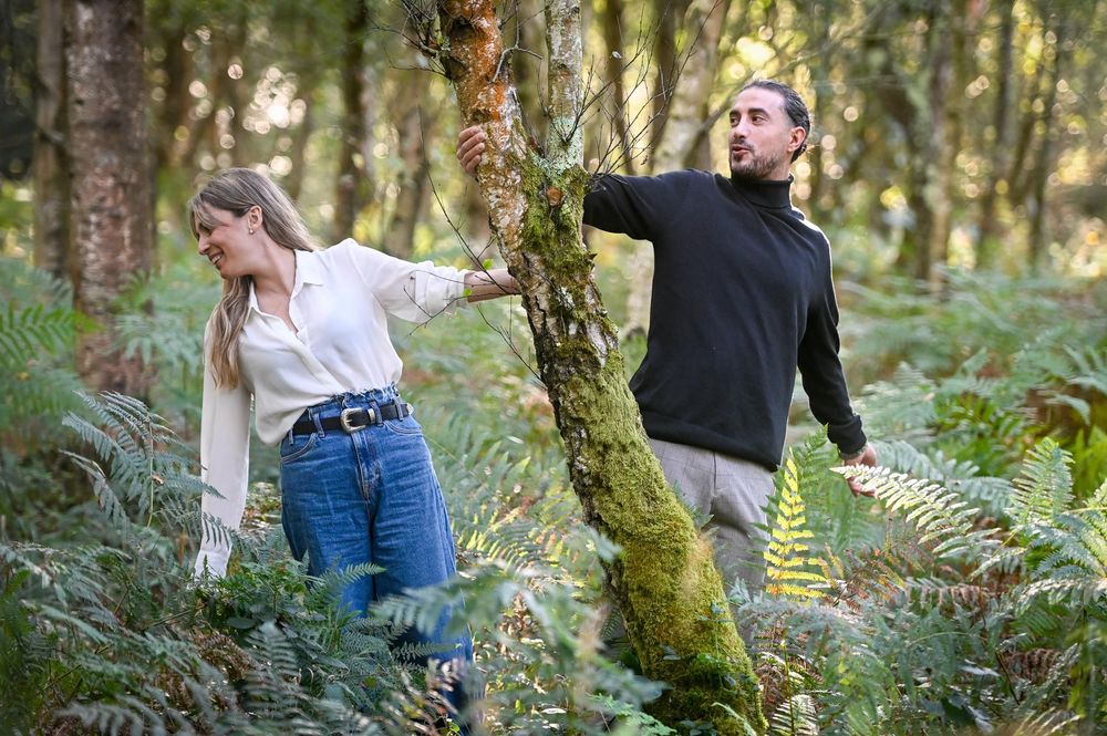 A couple walks together through a lush green forest with moss-covered trees and ferns in the background.