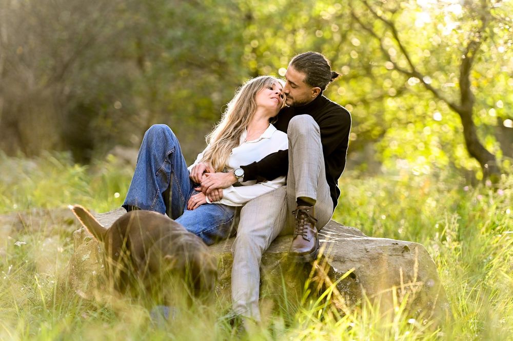 A romantic couple lounges together on a blanket in a sun-dappled meadow during a golden hour photoshoot.