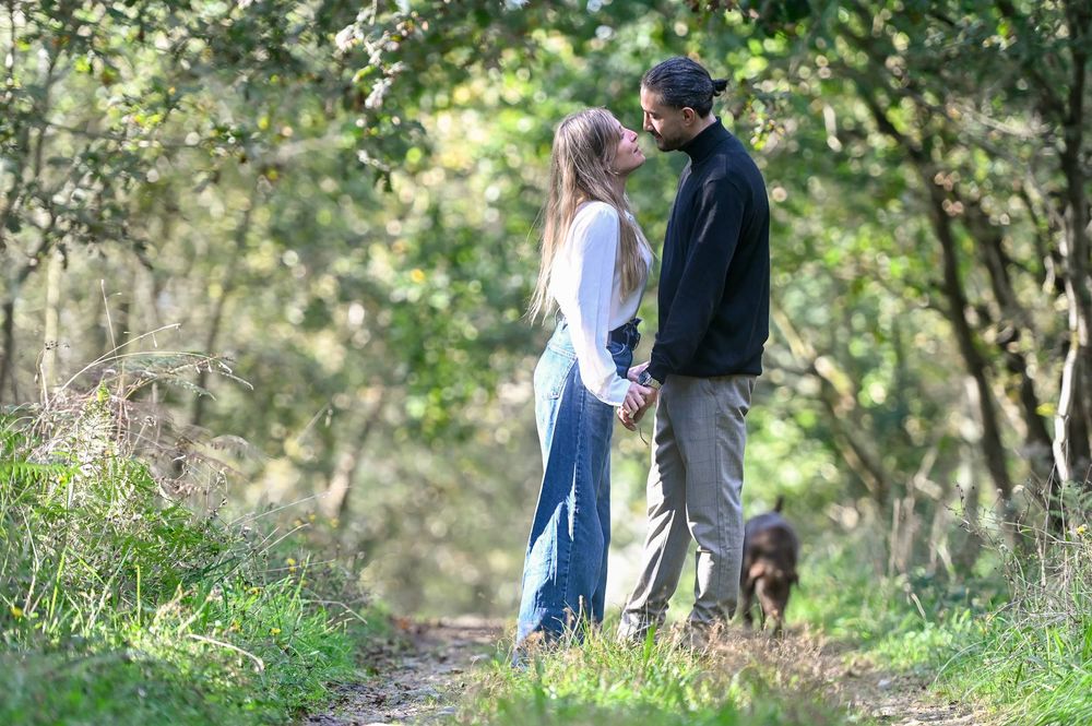 A couple shares a romantic moment on a wooded trail with their dog on a sunny afternoon.