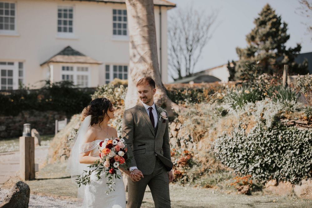 Bride holding a vibrant bouquet walks with groom through a sunlit garden at a country wedding.