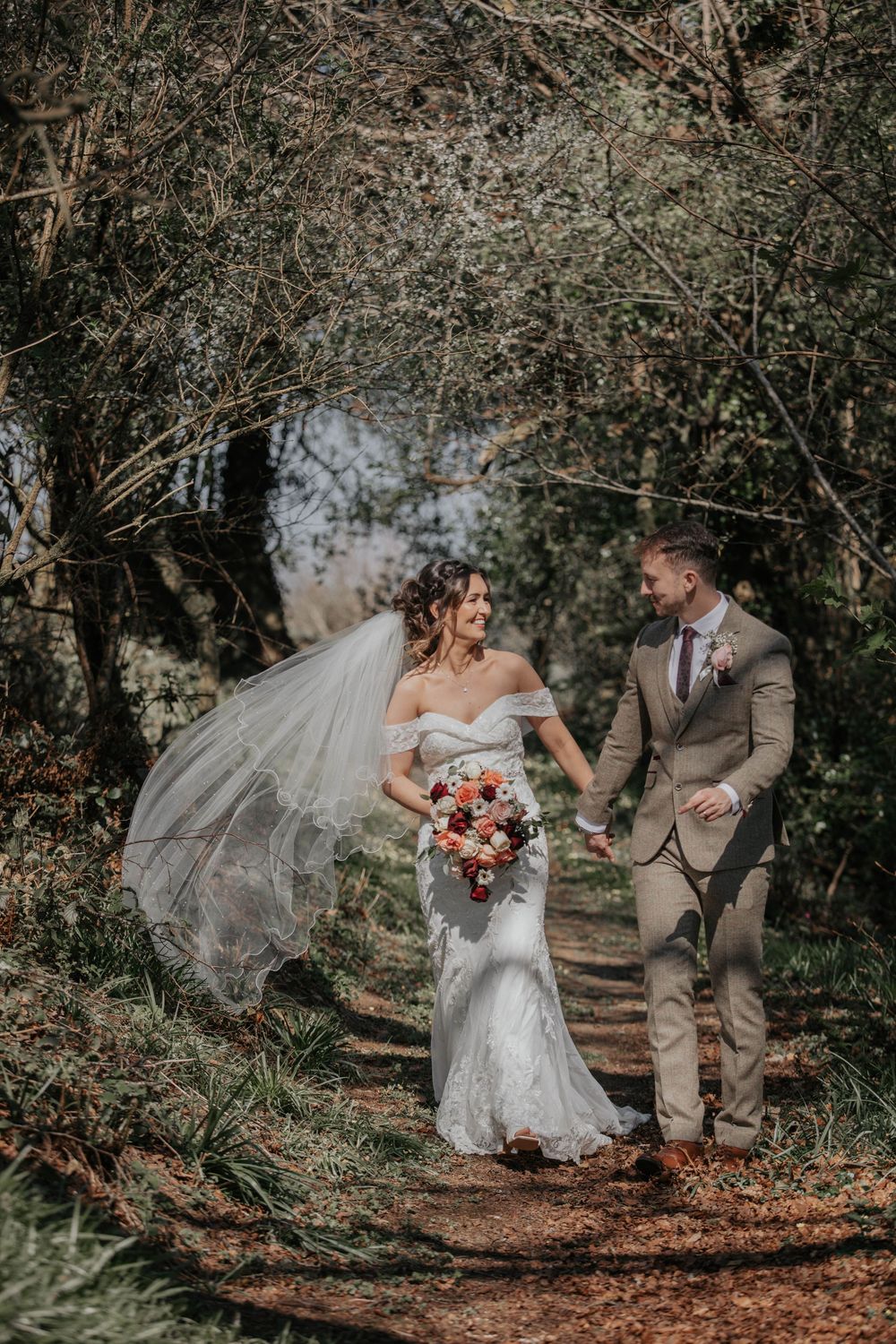 Bride and groom walking through a woodland path, veil flowing in the wind, holding a colorful bouquet.