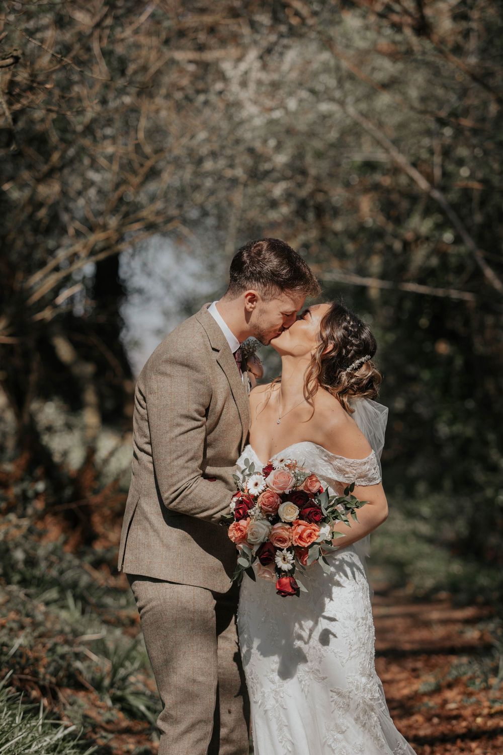 Bride and groom sharing a romantic kiss in a sunlit forest setting on their wedding day.