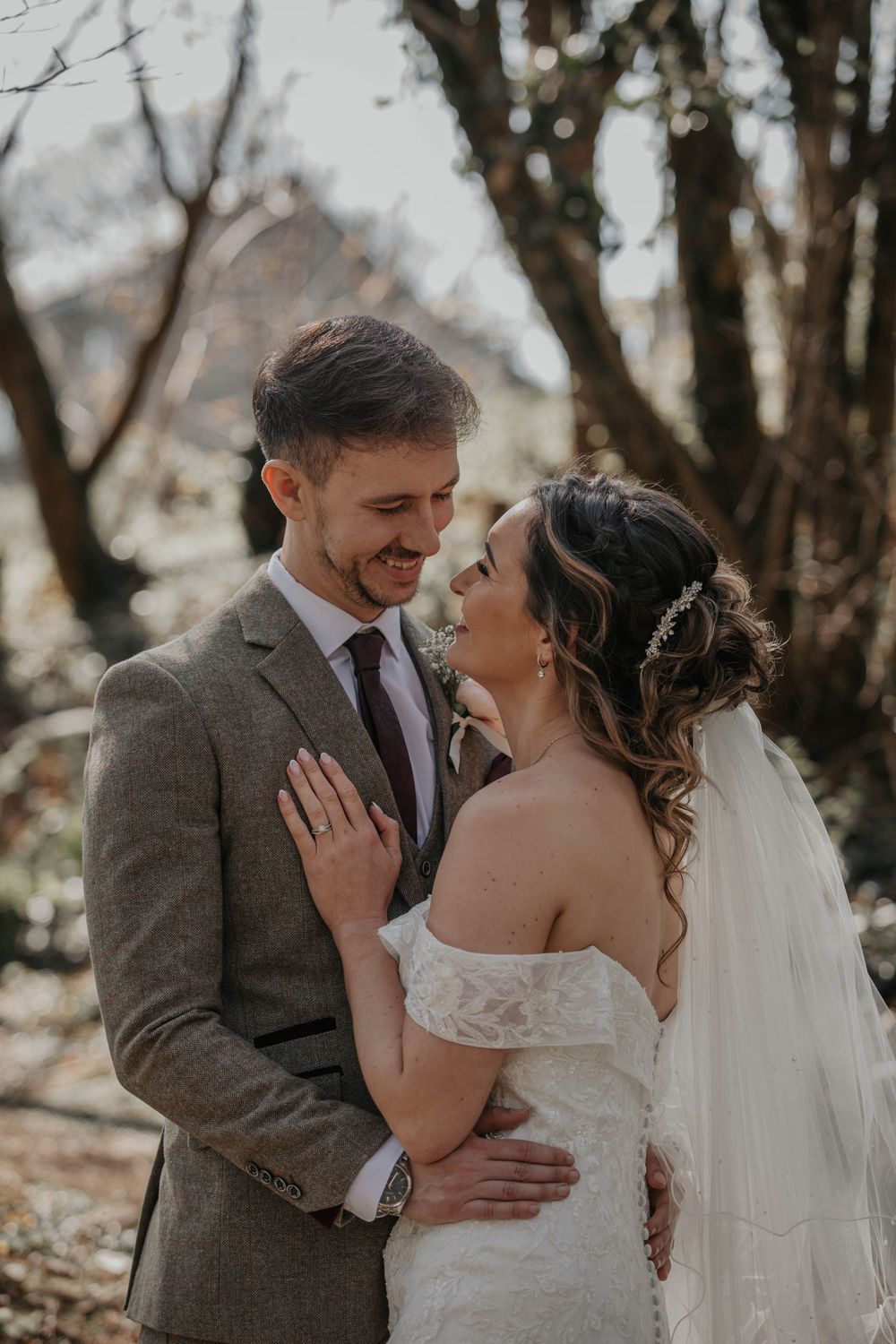 Smiling groom in grey suit embracing bride in off-shoulder lace wedding dress in woodland setting.