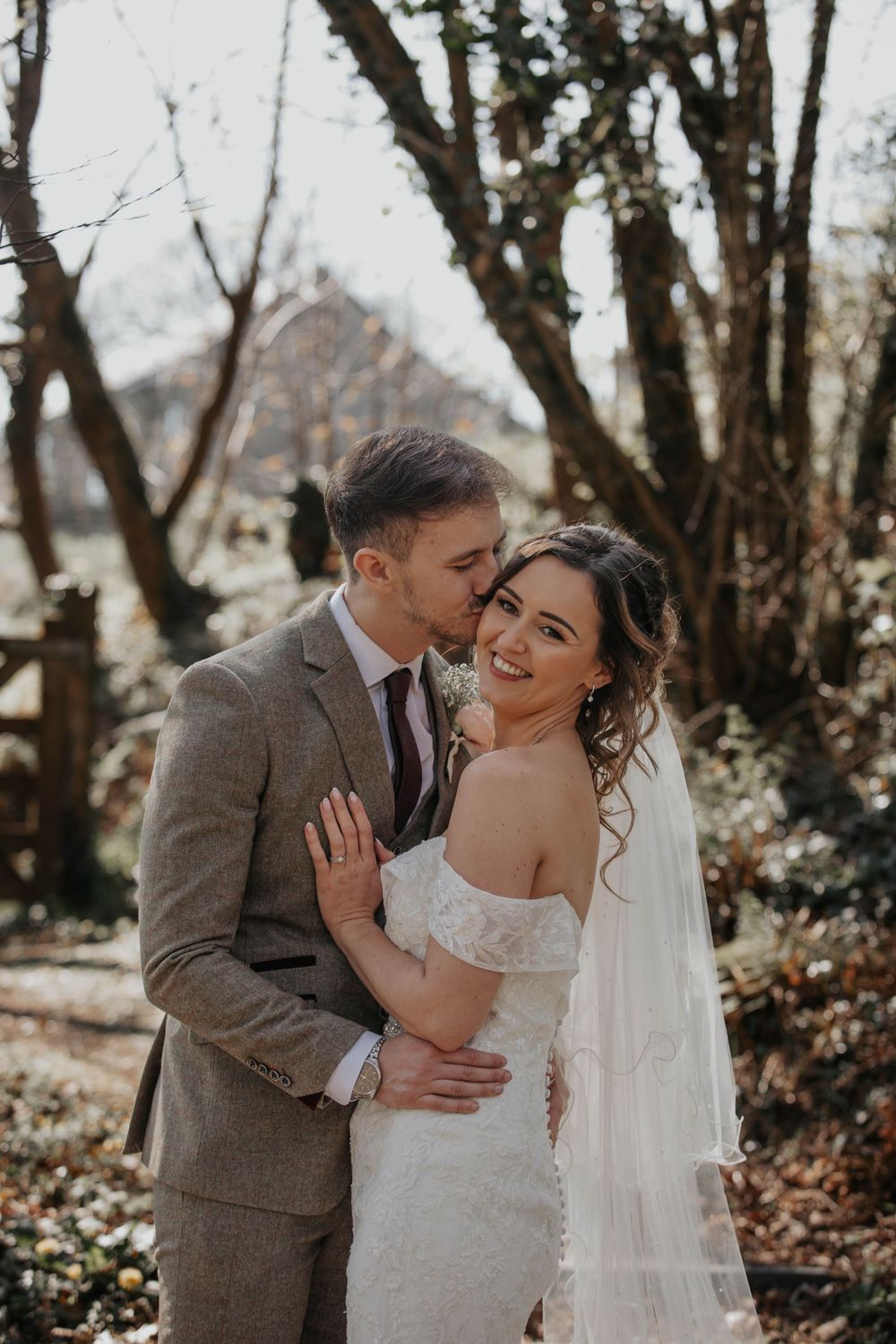 Groom kissing bride on cheek as she smiles in a lace wedding dress with veil outdoors.