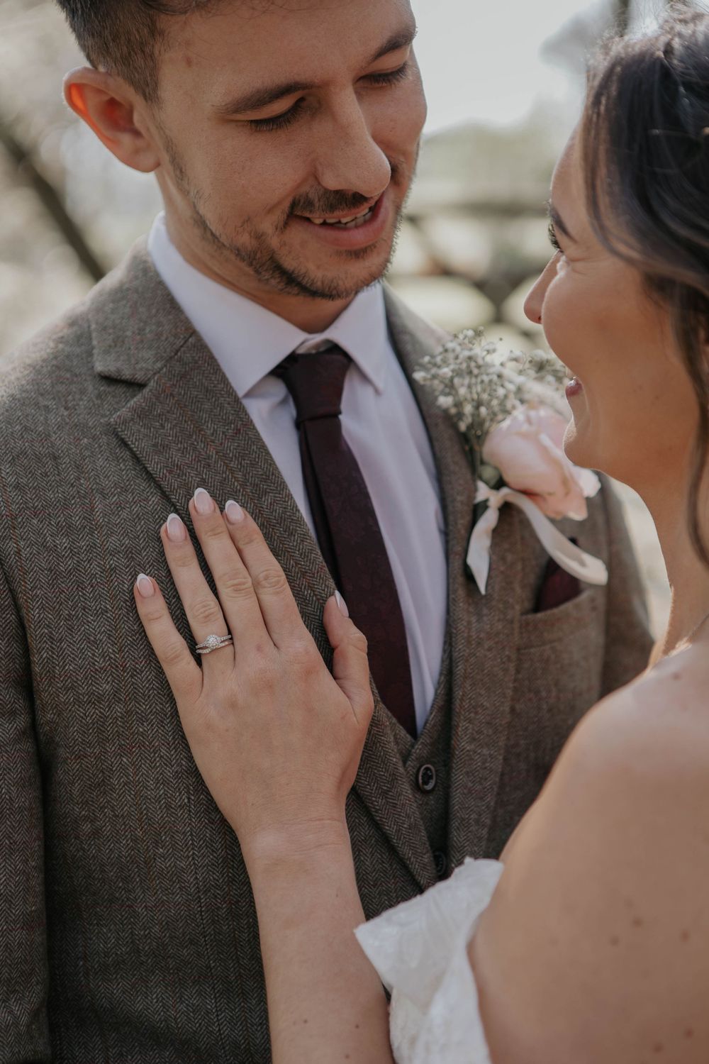 Close-up of bride showing wedding ring while groom smiles in grey herringbone suit with boutonniere.