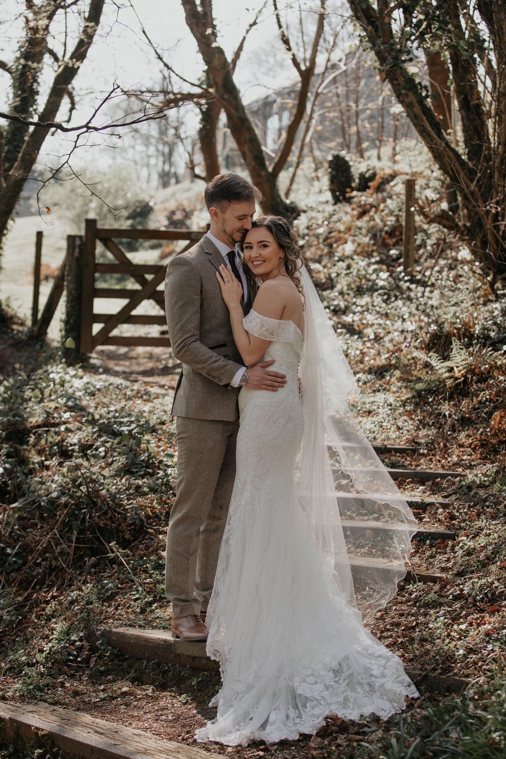 Bride and groom embracing near a wooden gate, brides lace veil flowing beautifully in natural light.