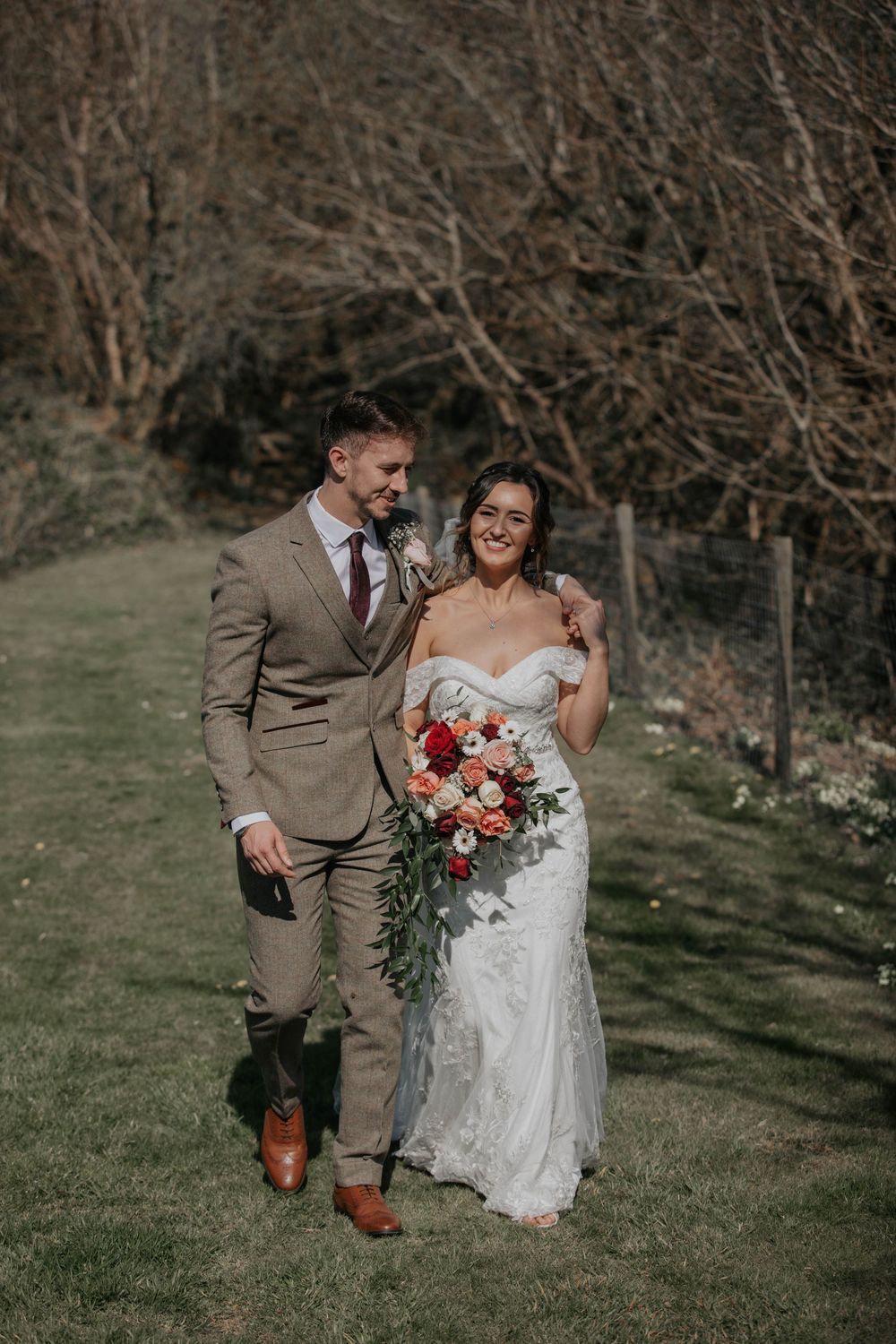 Bride and groom walking together on sunny lawn, bride holding vibrant red and orange bridal bouquet.