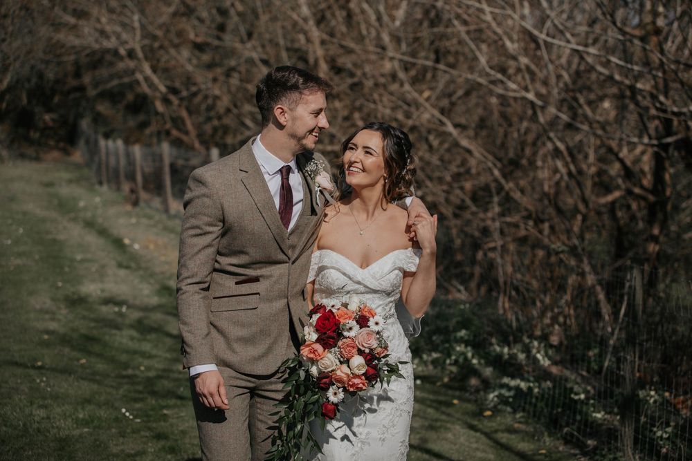 Bride and groom smiling at each other outdoors, bride holding a lush red and white floral bouquet.