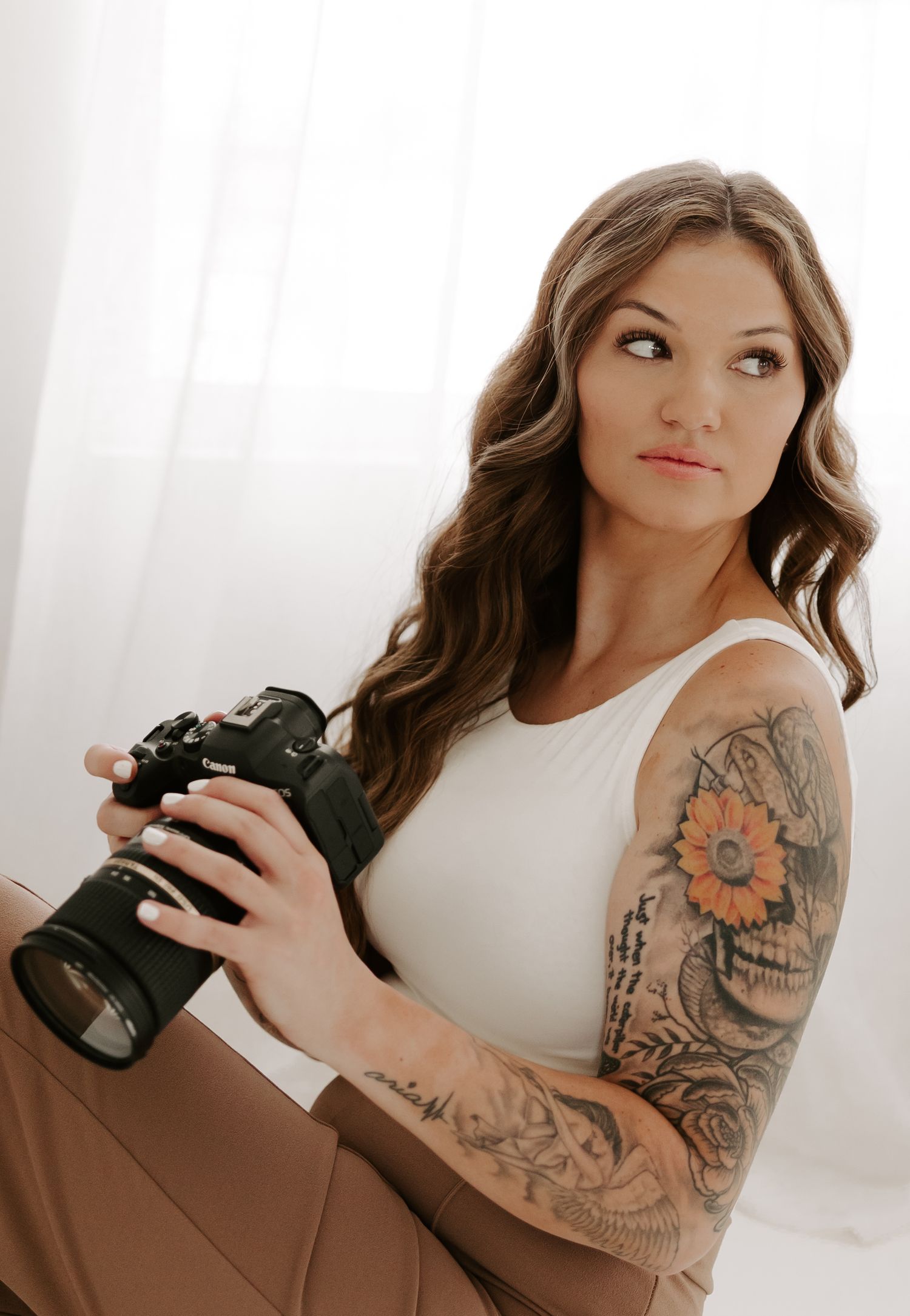 A photographer with floral arm tattoos holding a professional camera in a studio setting.