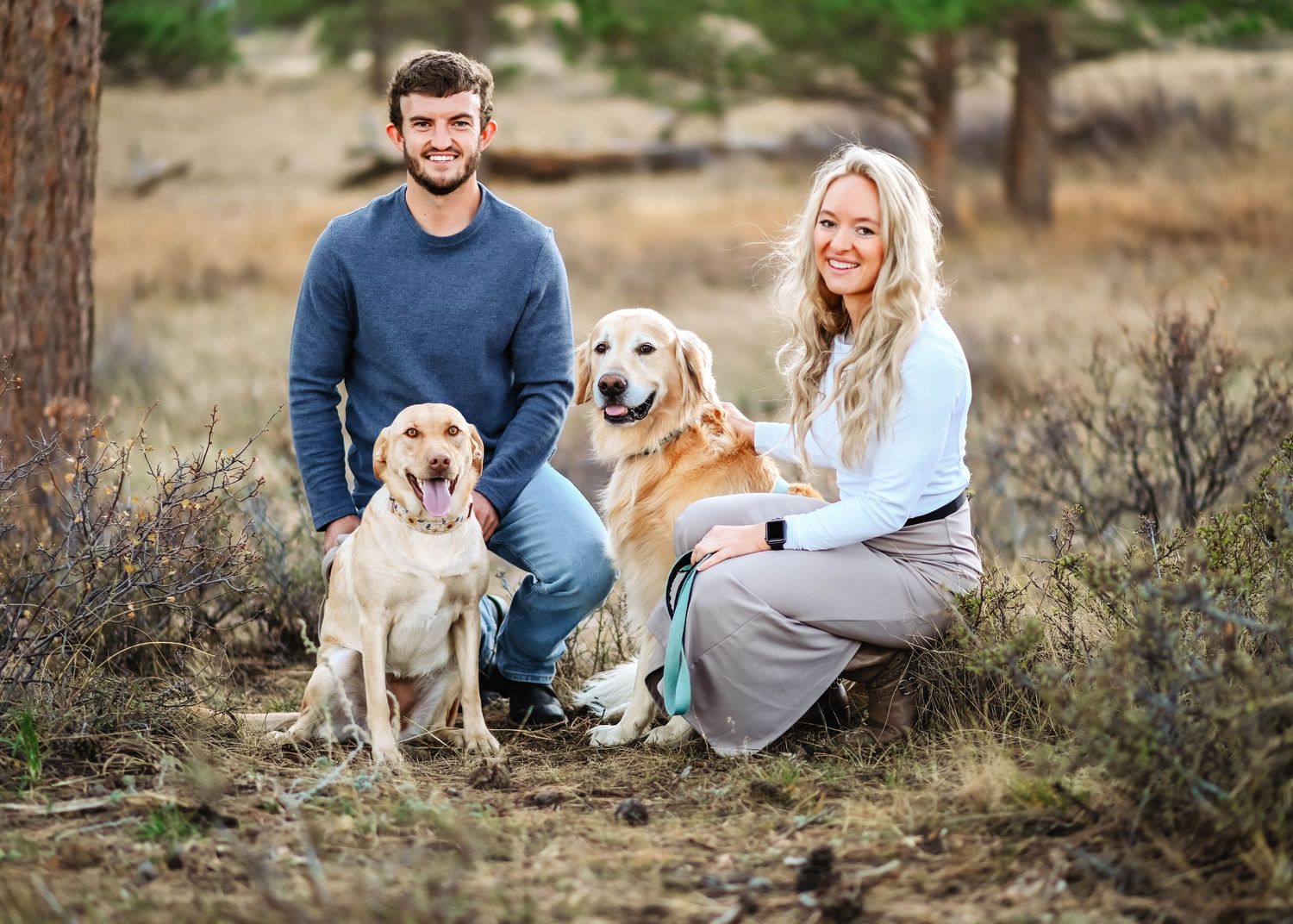 A couple sits outdoors with their two golden retriever dogs during a casual photo session in a natural setting.
