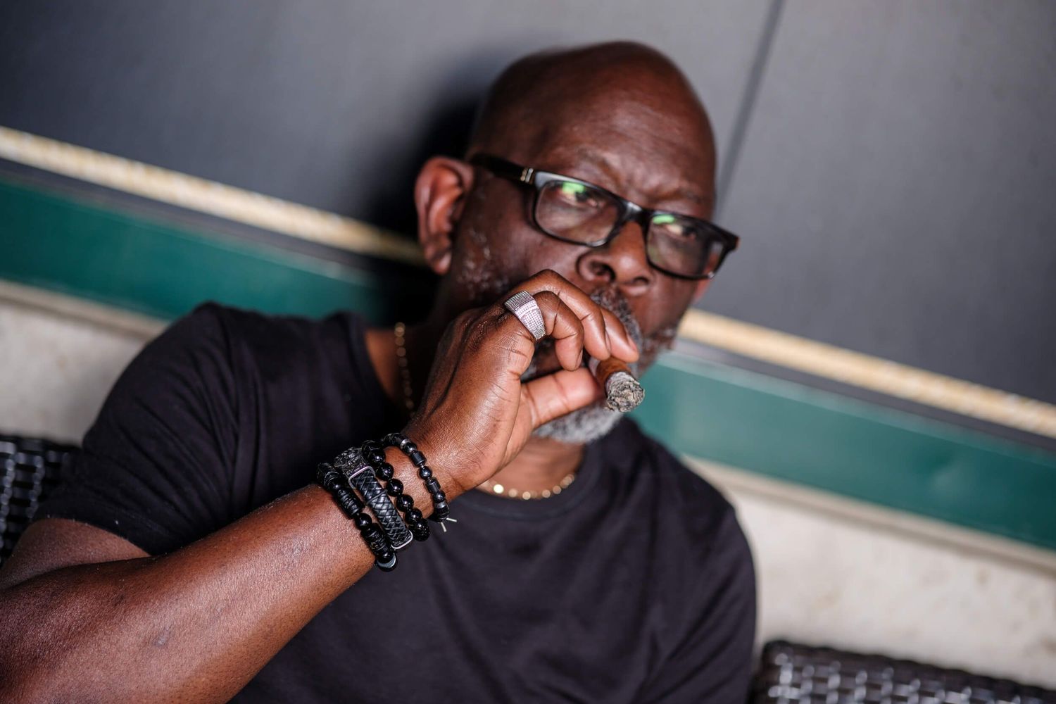 A person wearing glasses sits in contemplation with their hand near their face in a dark setting.