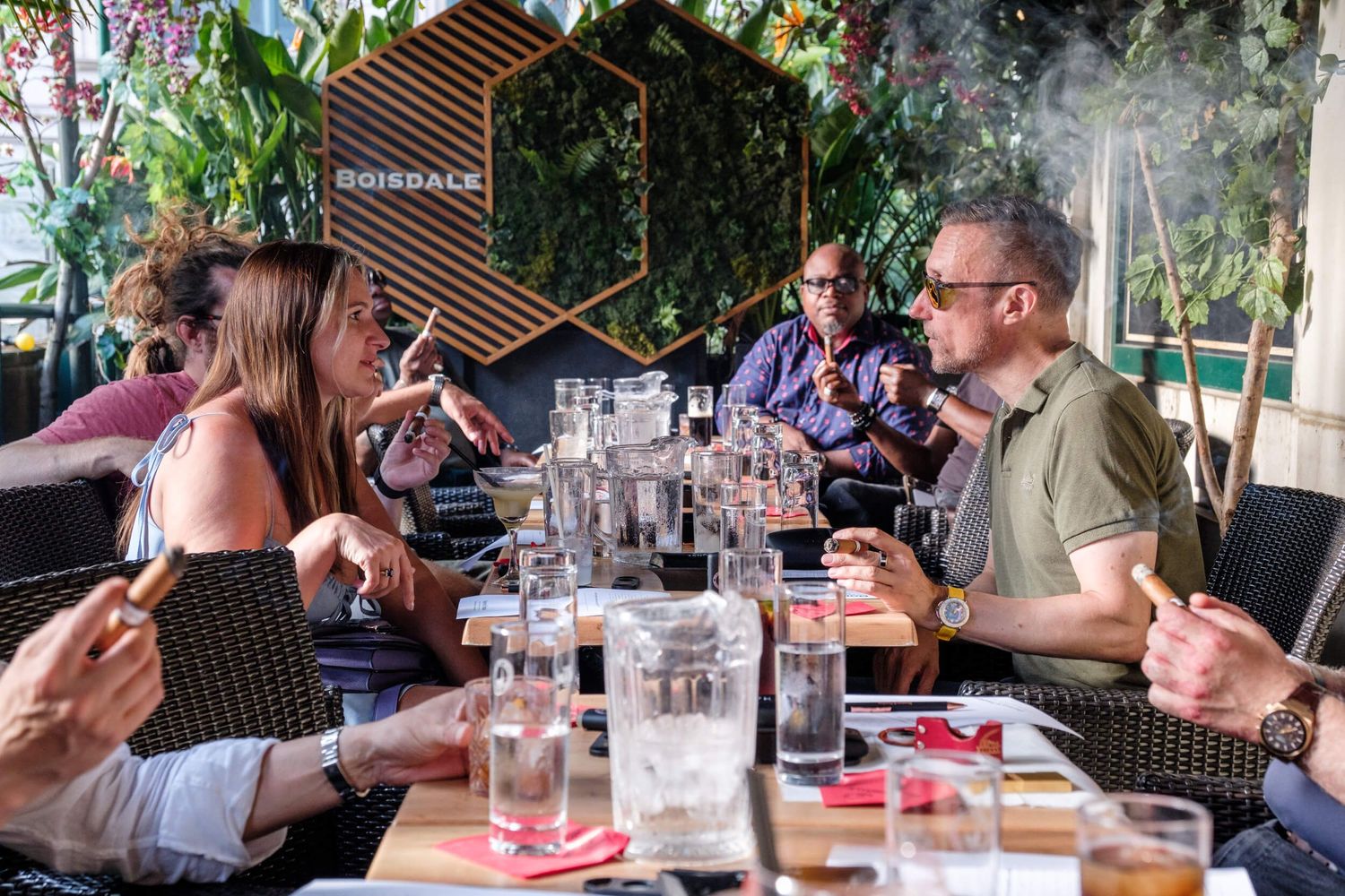 People enjoying drinks and conversation at an outdoor restaurant patio with lush greenery in the background.