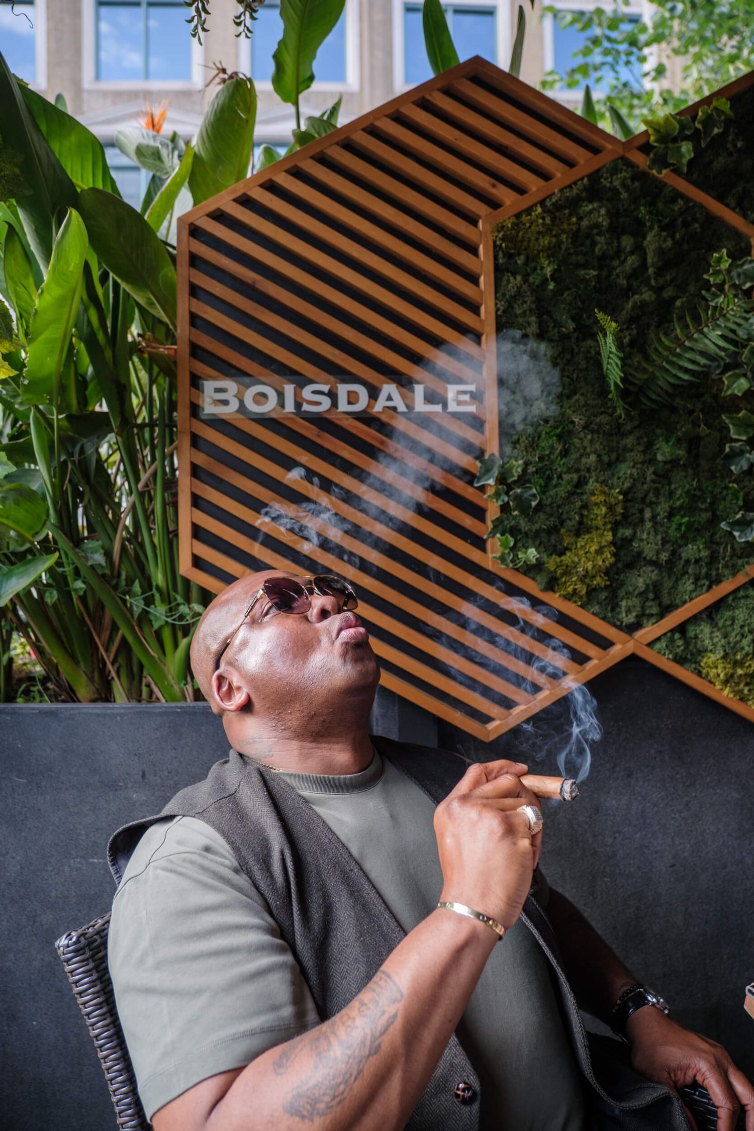 A person relaxes in a modern outdoor patio space with wooden slat details and tropical plants in the background.
