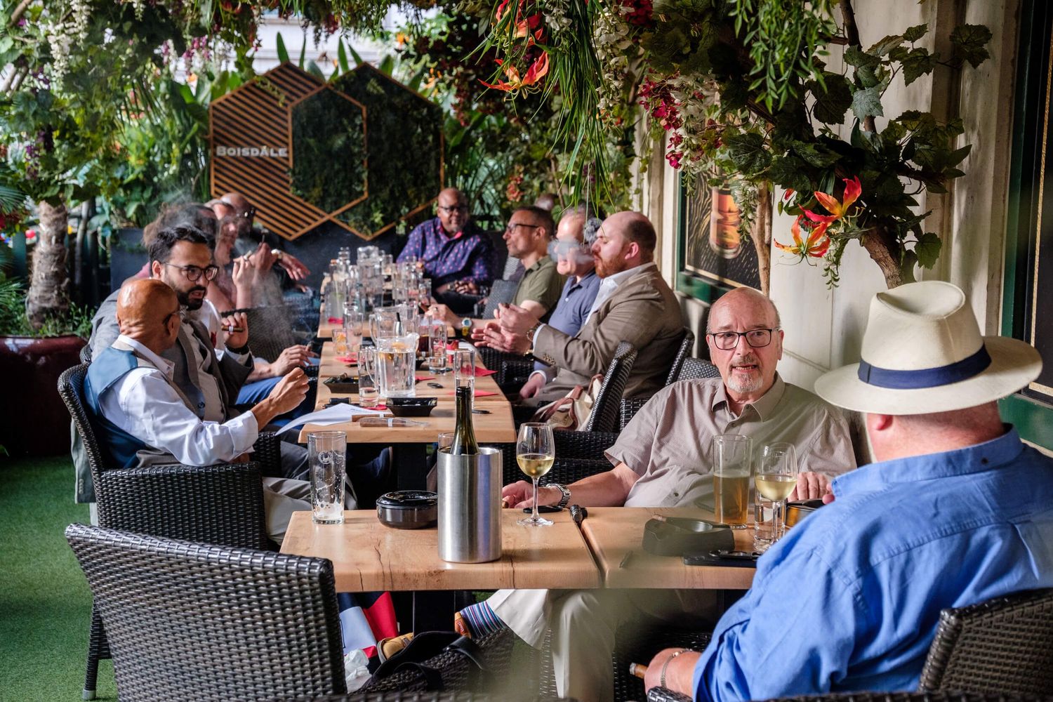 Outdoor restaurant patio filled with diners enjoying a meal under string lights and greenery on a sunny day.