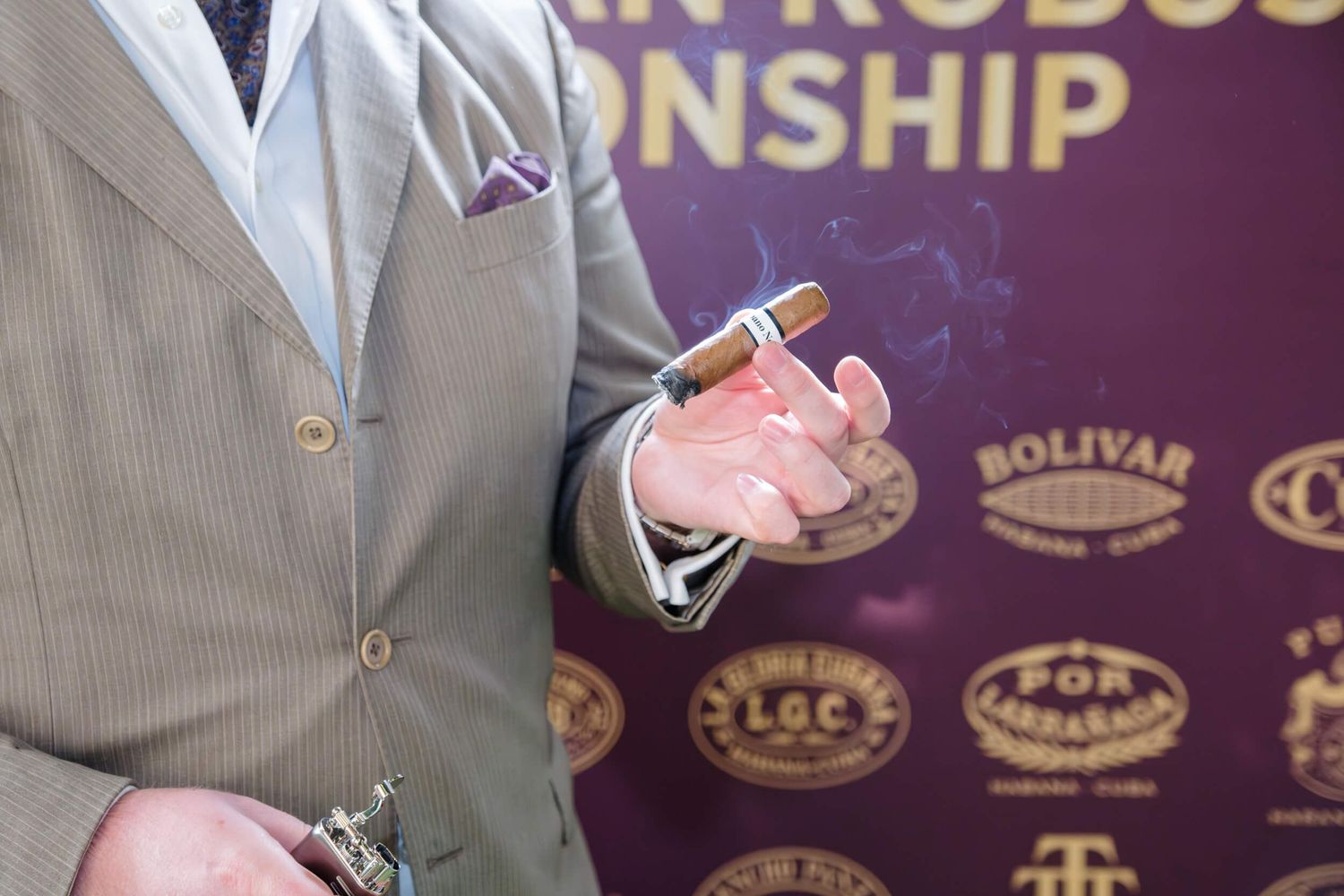 A person in a tan suit jacket holds a pink cigar lighter against a purple backdrop with brand logos.