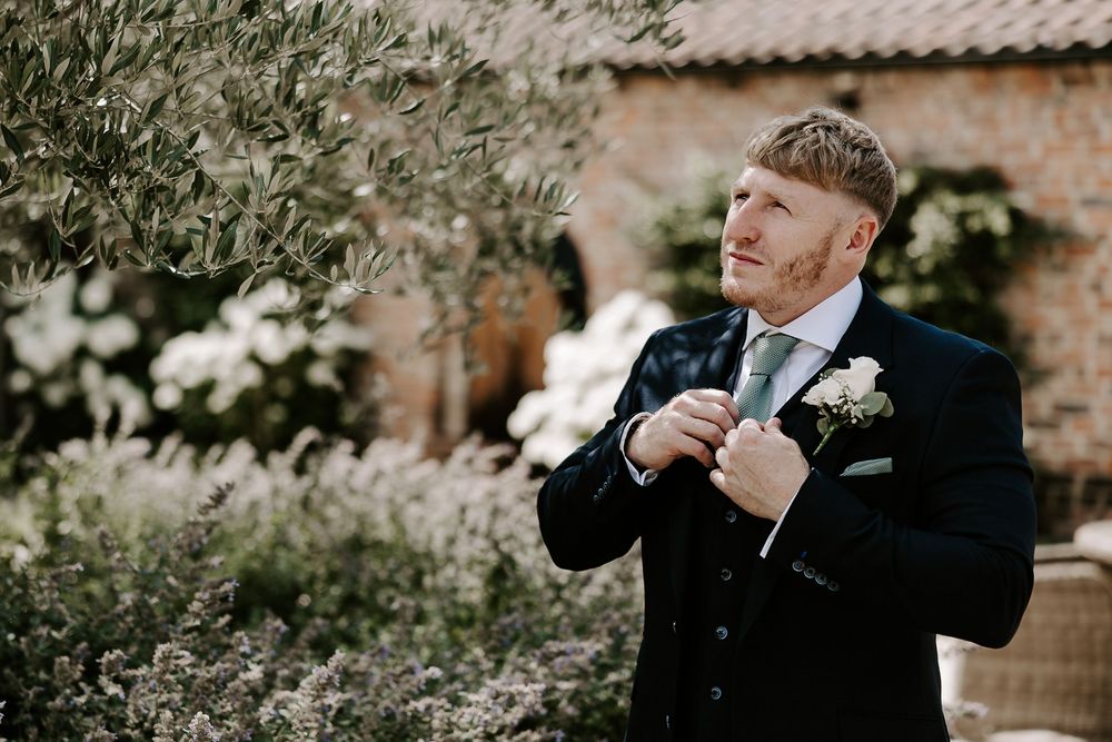 Candid moments of someone in a dark suit adjusting their tie outdoors against a garden backdrop.