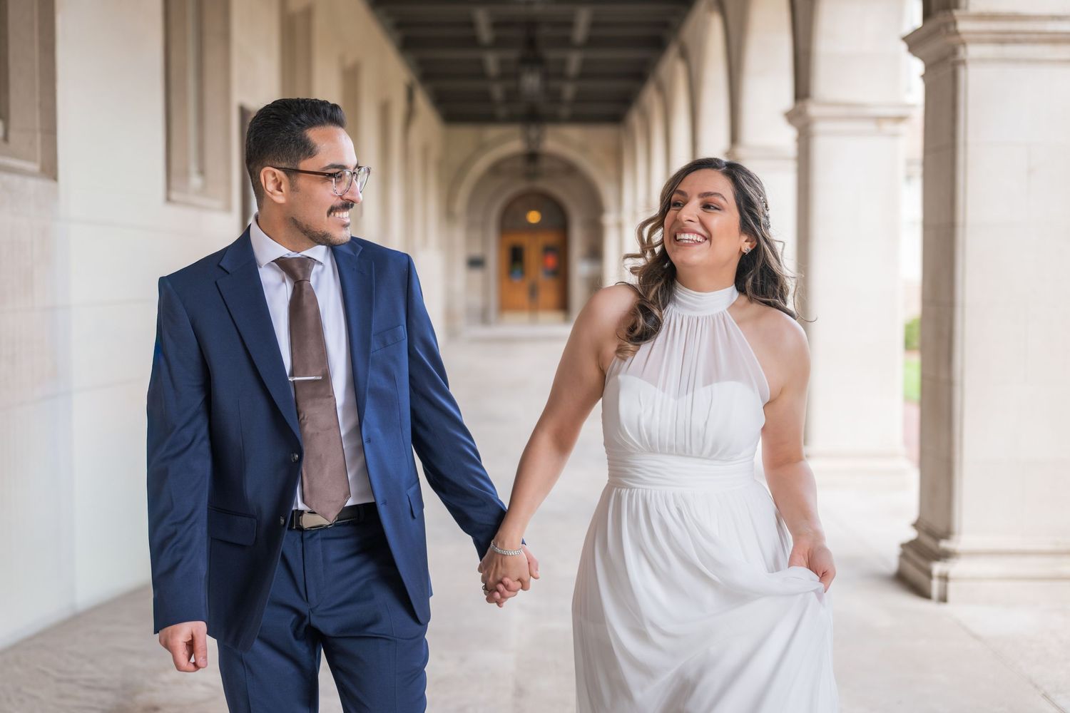 A couple in wedding attire laughs together while walking through an elegant columned hallway.