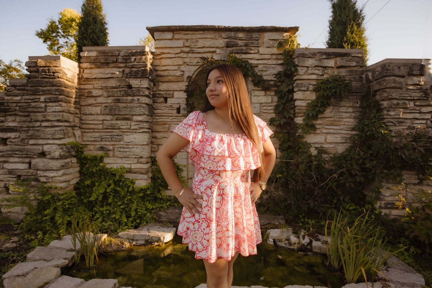 A person in a pink floral dress poses in front of a stone wall during golden hour.
