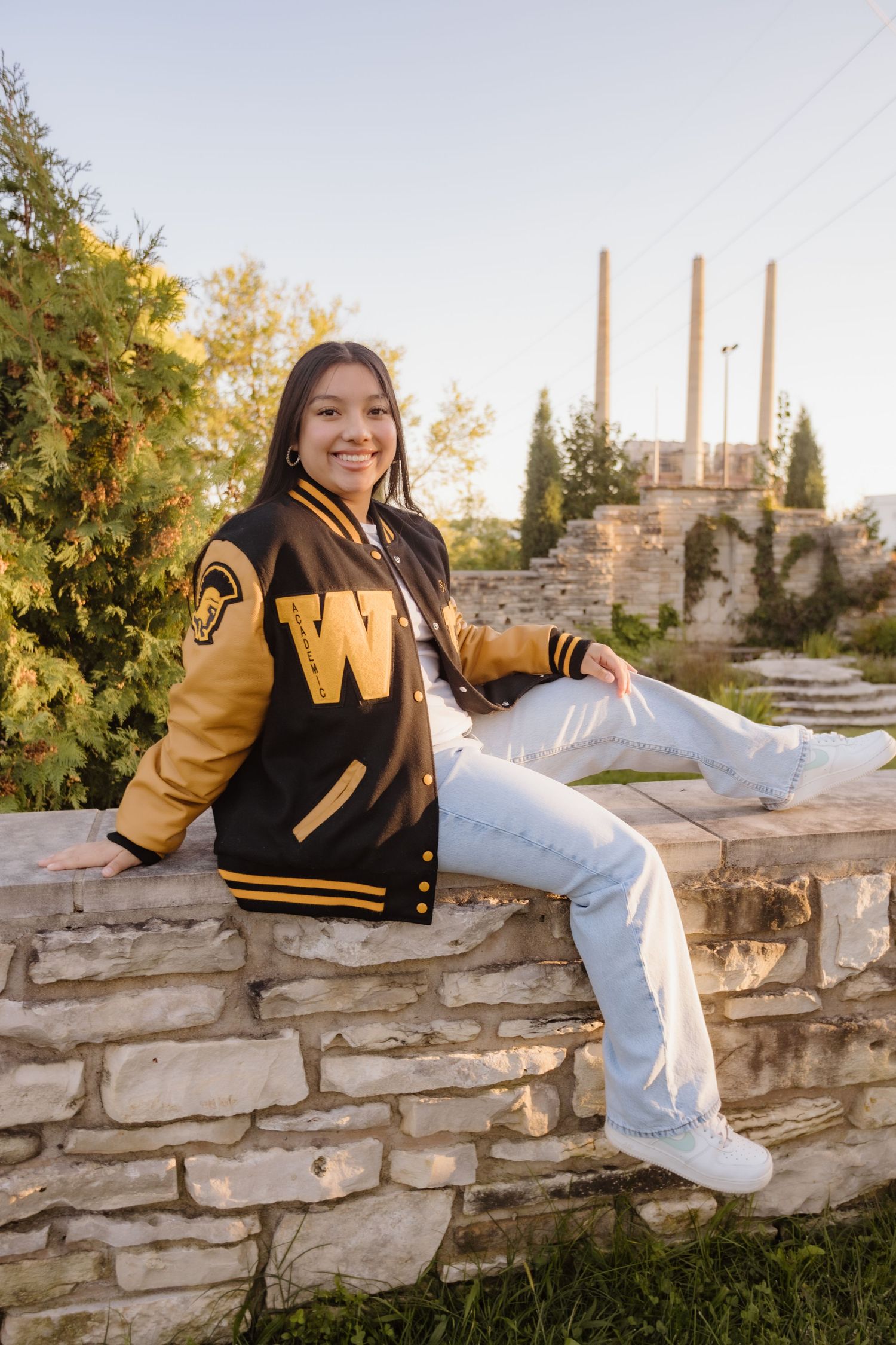 A person in a black and gold varsity jacket poses casually on a stone wall during golden hour sunset.