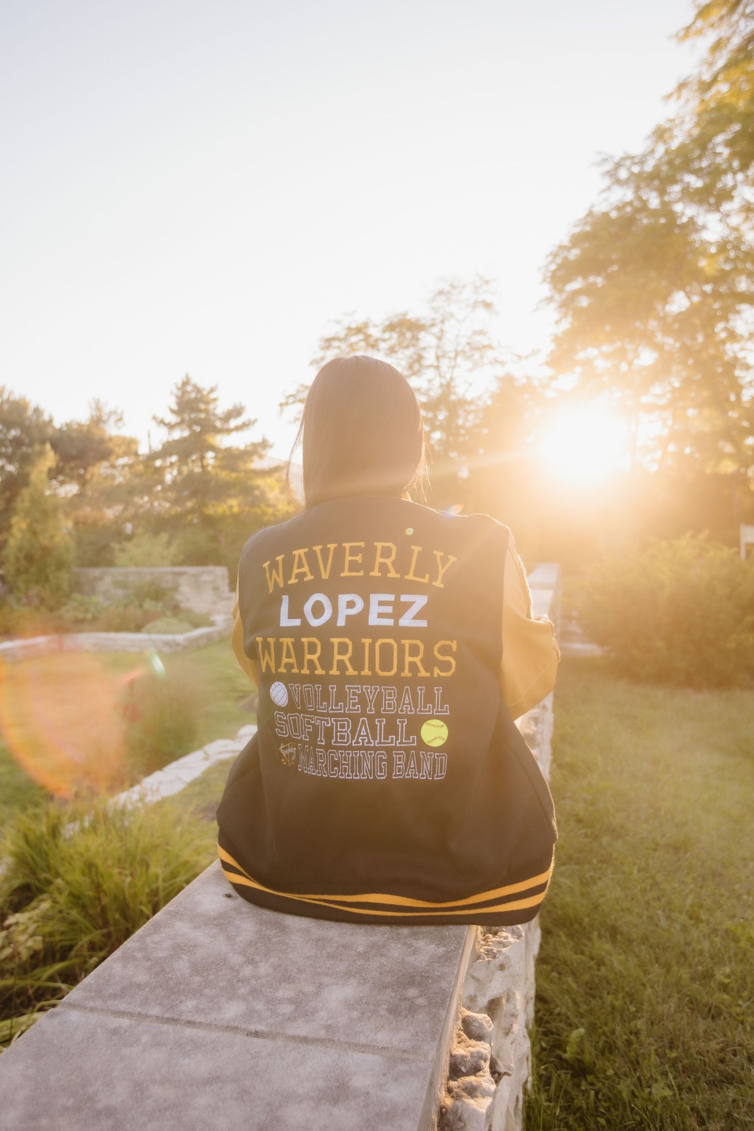 Person wearing a Waverly Lopez Warriors varsity jacket sits on concrete wall at sunset with sun flare in background.