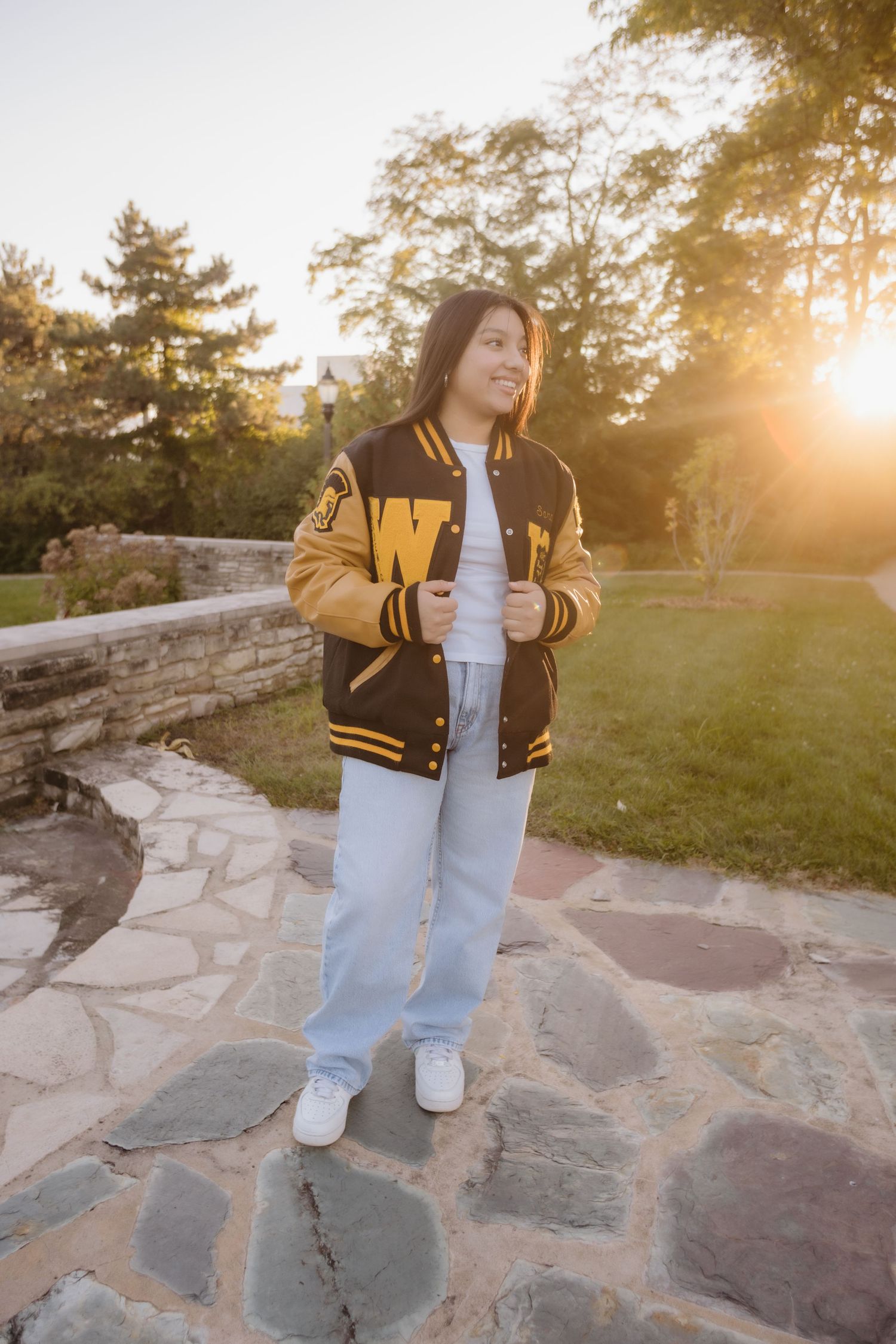 A figure in a varsity jacket and light jeans stands on stone steps during golden hour sunset.