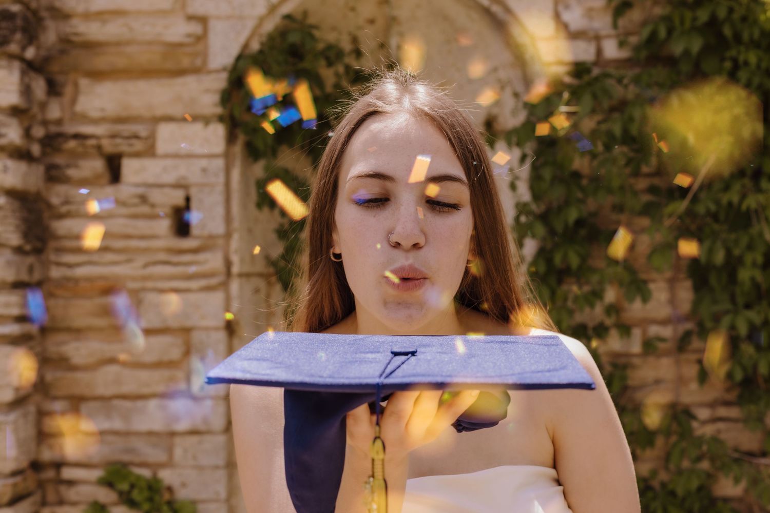A bride in a white dress holds a book while reading outdoors against a stone wall with string lights and greenery.
