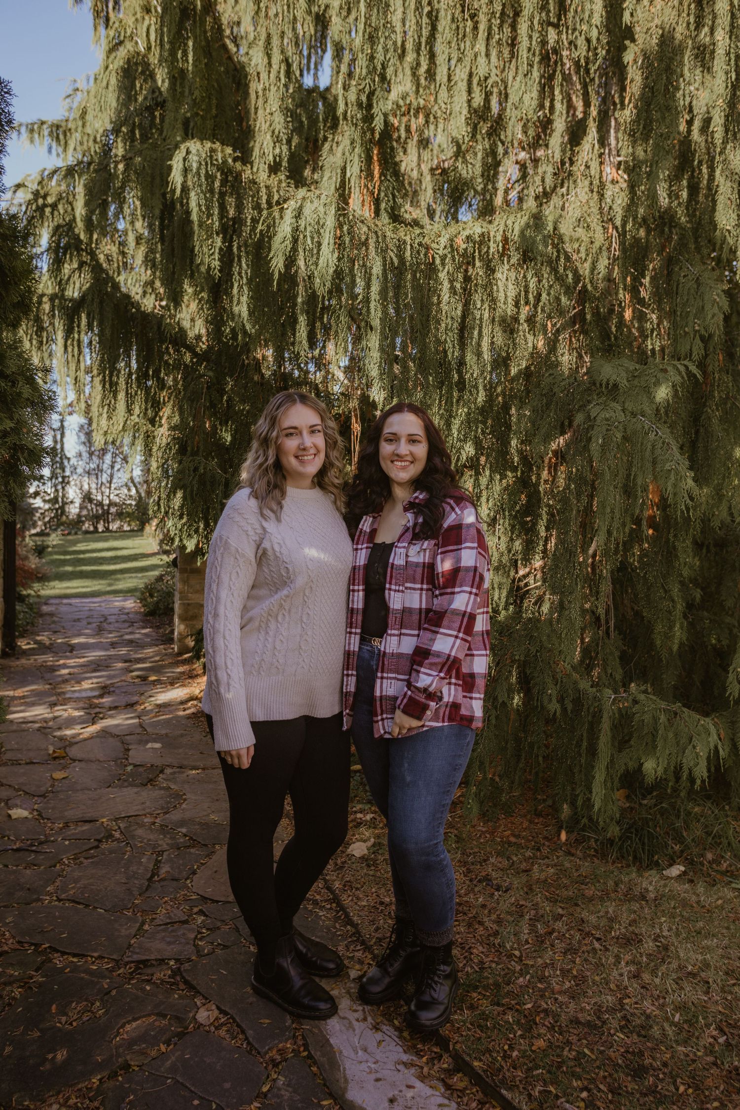 Two people pose together under a dramatic weeping willow tree on an autumn day.