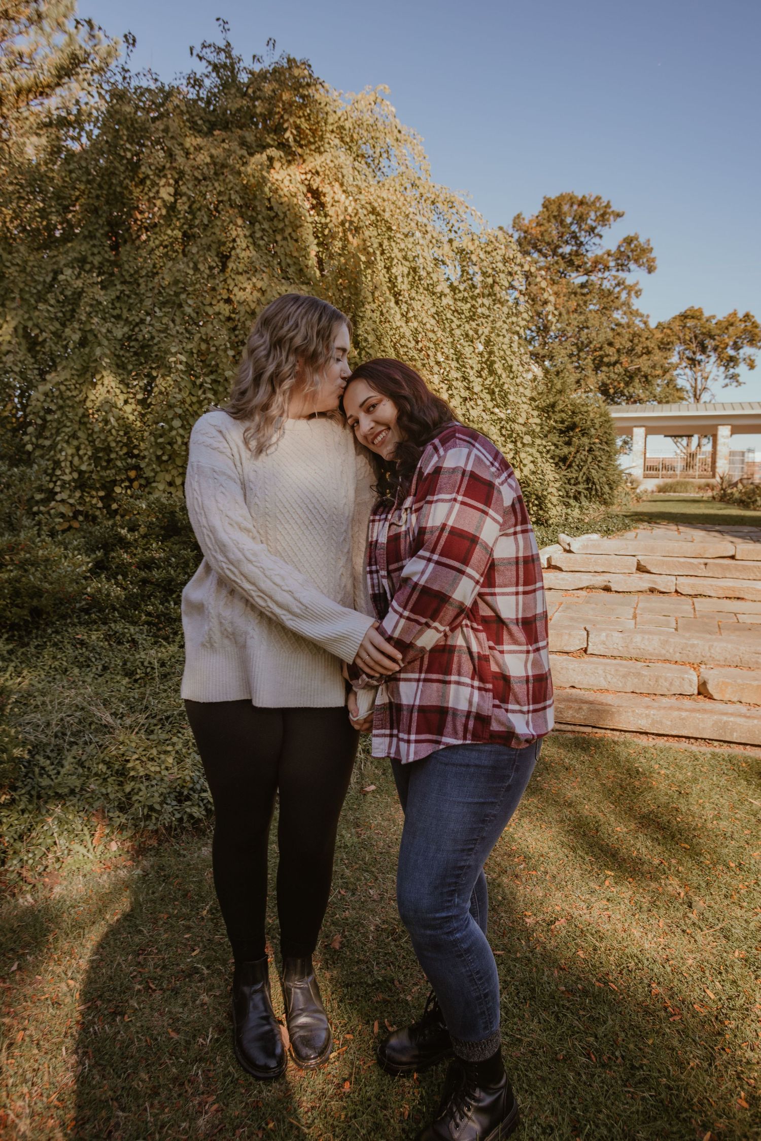 A couple embraces outdoors on a sunny autumn day with golden foliage and stone wall in the background.