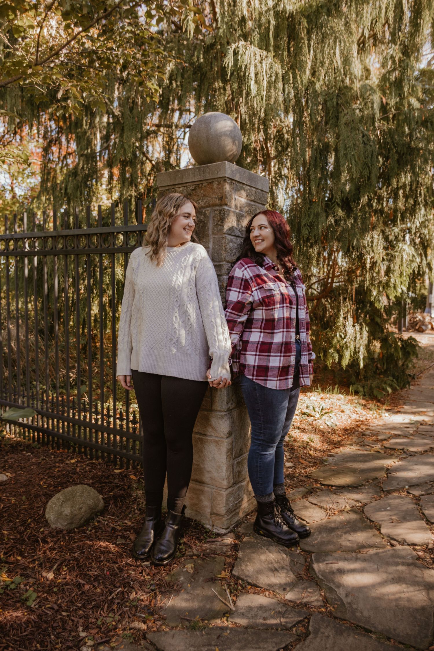 Two people stand together on a stone path surrounded by autumn foliage and a rustic fence in soft natural lighting.