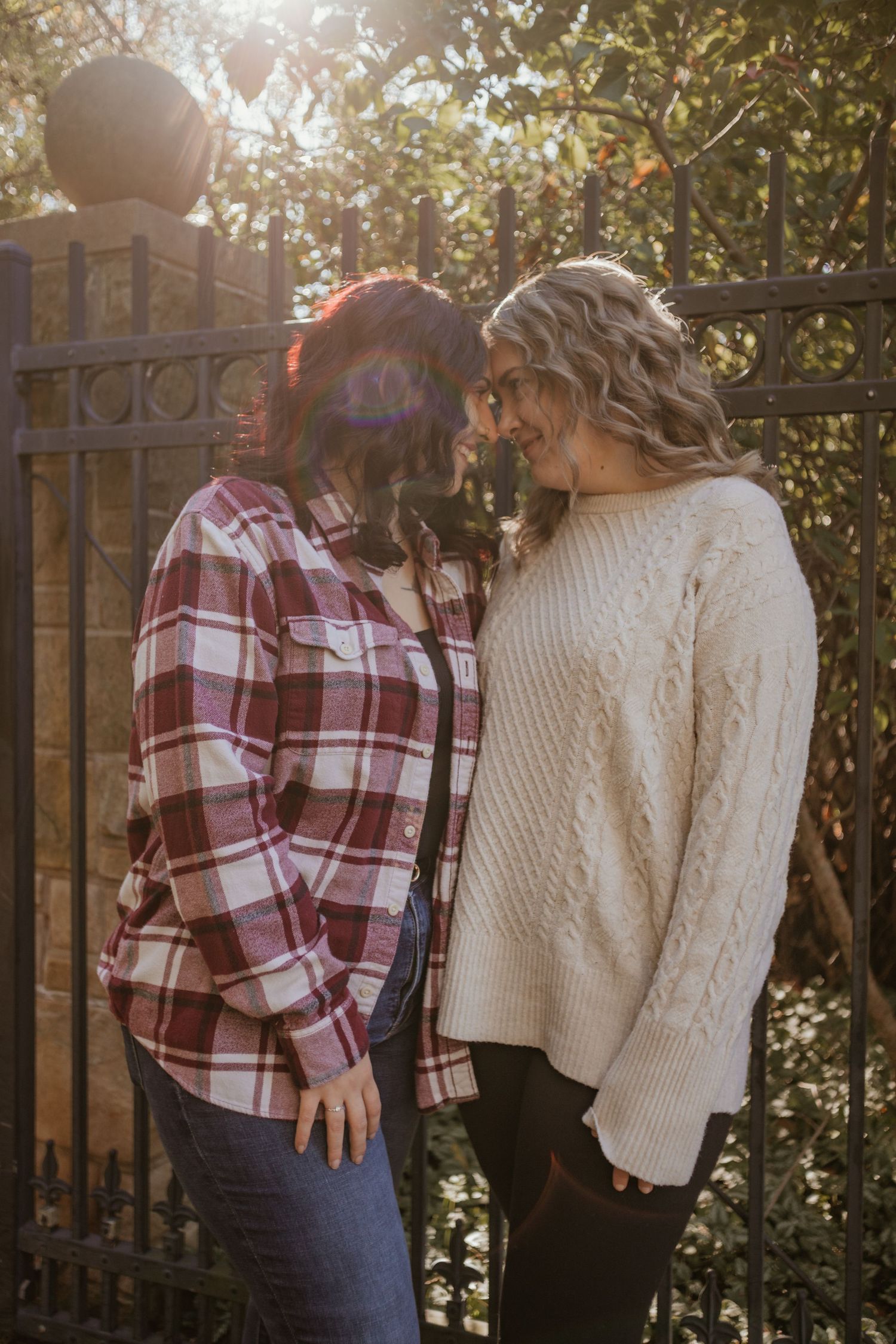 Two people wearing cozy autumn outfits embrace outdoors against a wooden fence in warm sunlight.