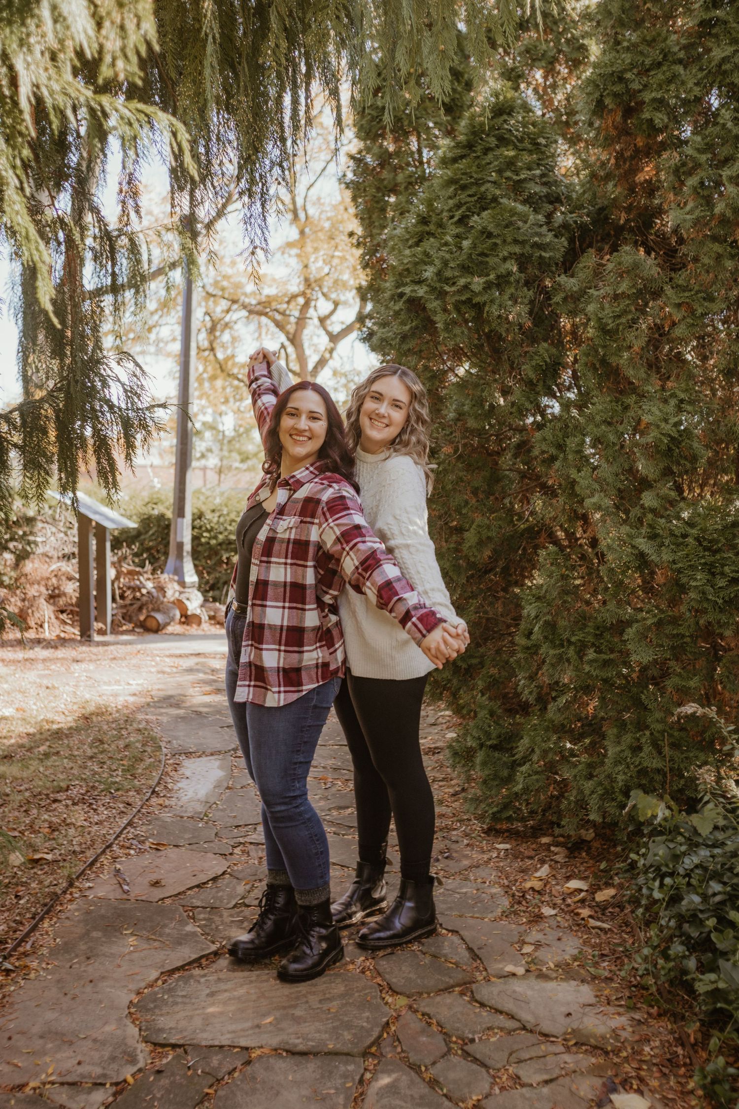 Two people in coordinating plaid and cream outfits pose together on a stone path surrounded by evergreen trees.
