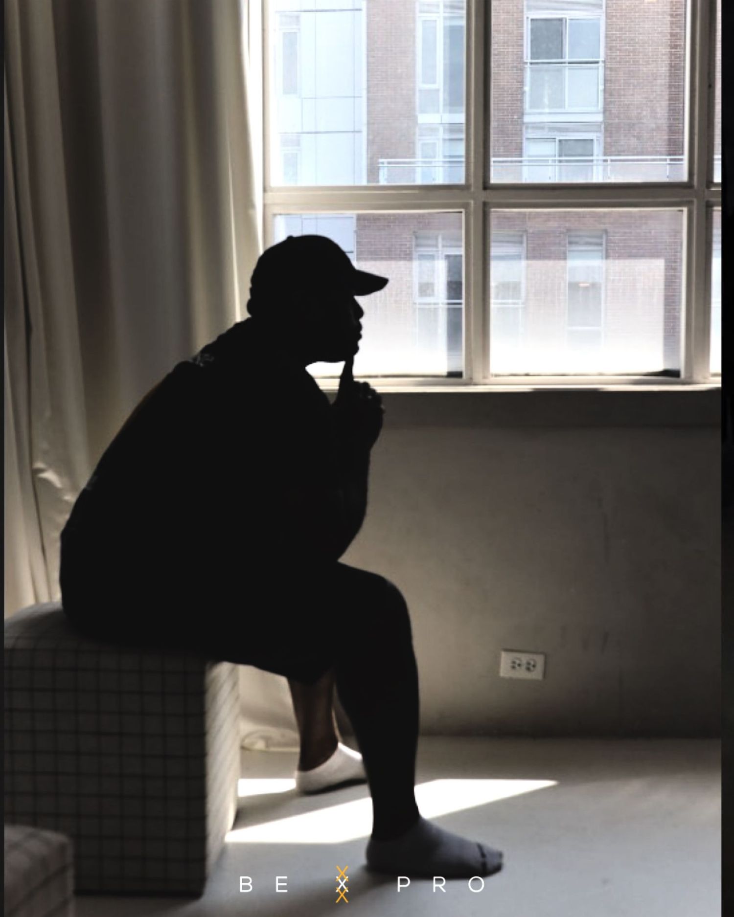 A dramatic silhouette of a person in a cap sitting pensively by a bright window in an empty room.