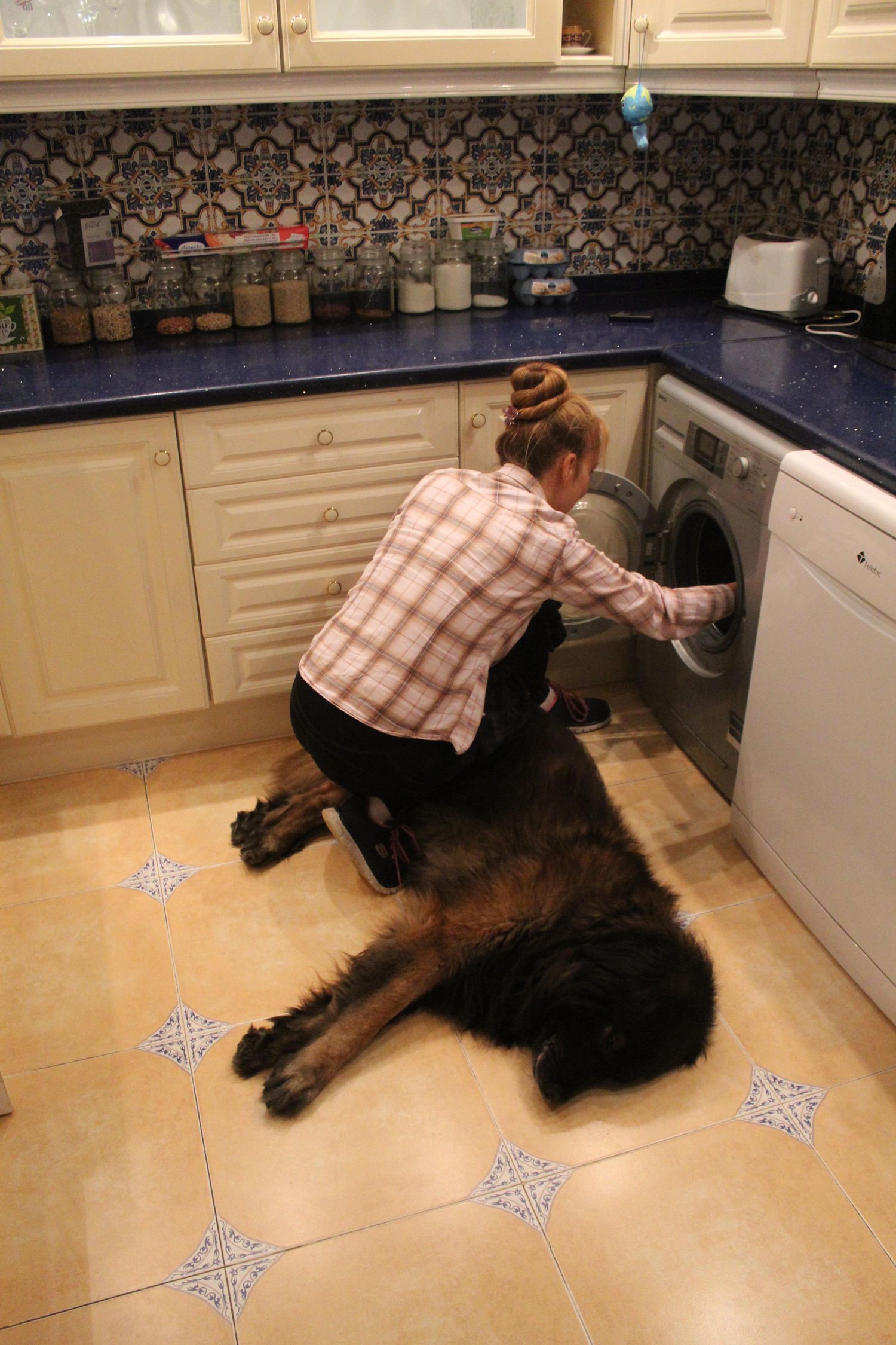 Woman crouching by washing machine in kitchen while large long-haired black and tan dog sits beside her.