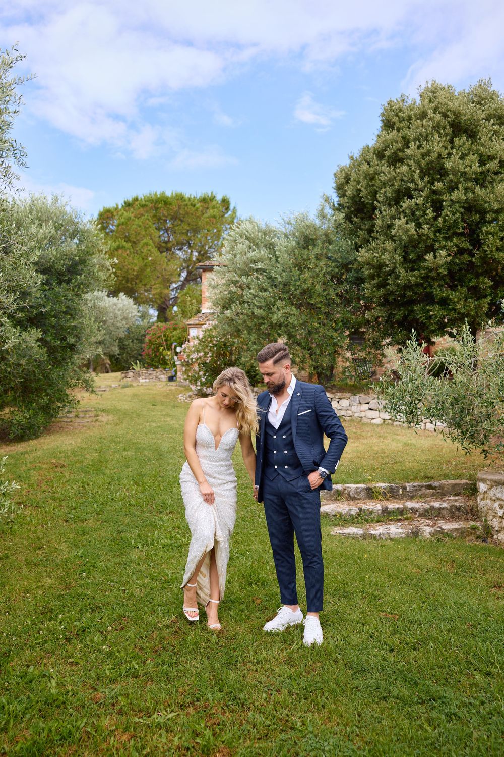 Bride and groom walk hand in hand through a lush green garden with historic stone ruins in the background.