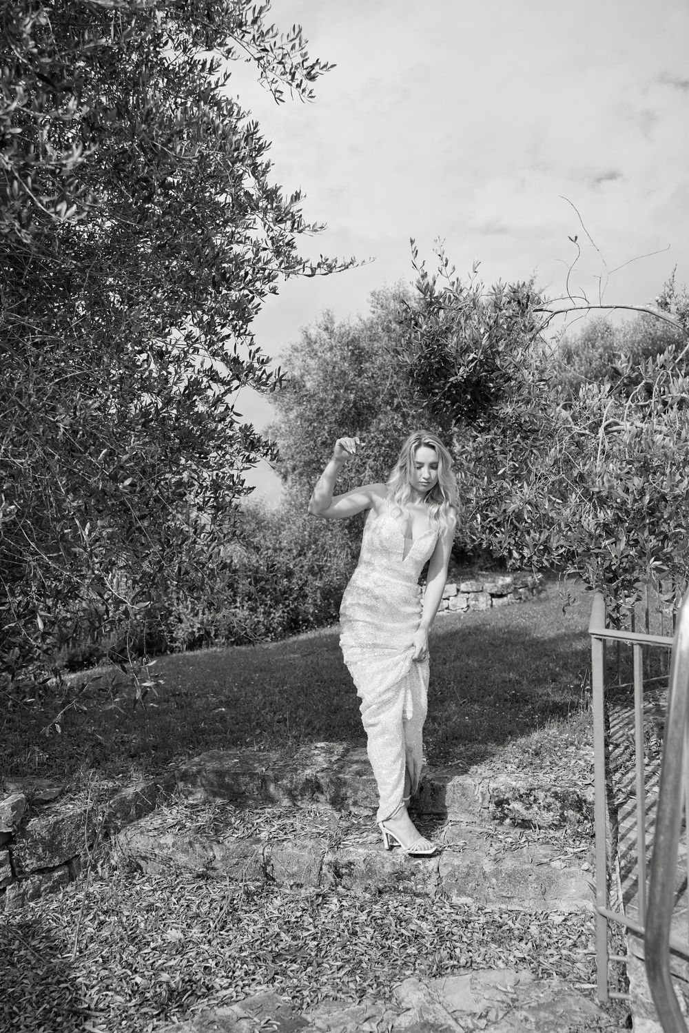 Elegant bride in white gown poses on stone steps surrounded by lush olive trees in a garden setting.