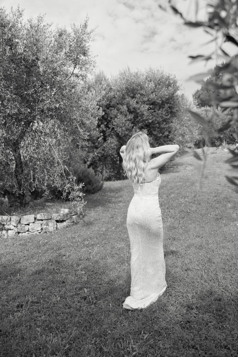 Bride in white lace gown standing in lush garden with stone wall and trees.