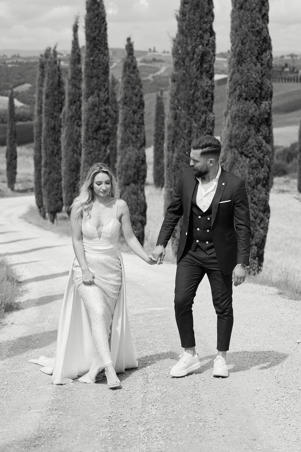 Bride and groom walking hand in hand along cypress tree lined Tuscan road.