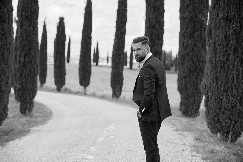 Elegant man in black suit standing on a tree-lined gravel path in Tuscany, black and white photo.