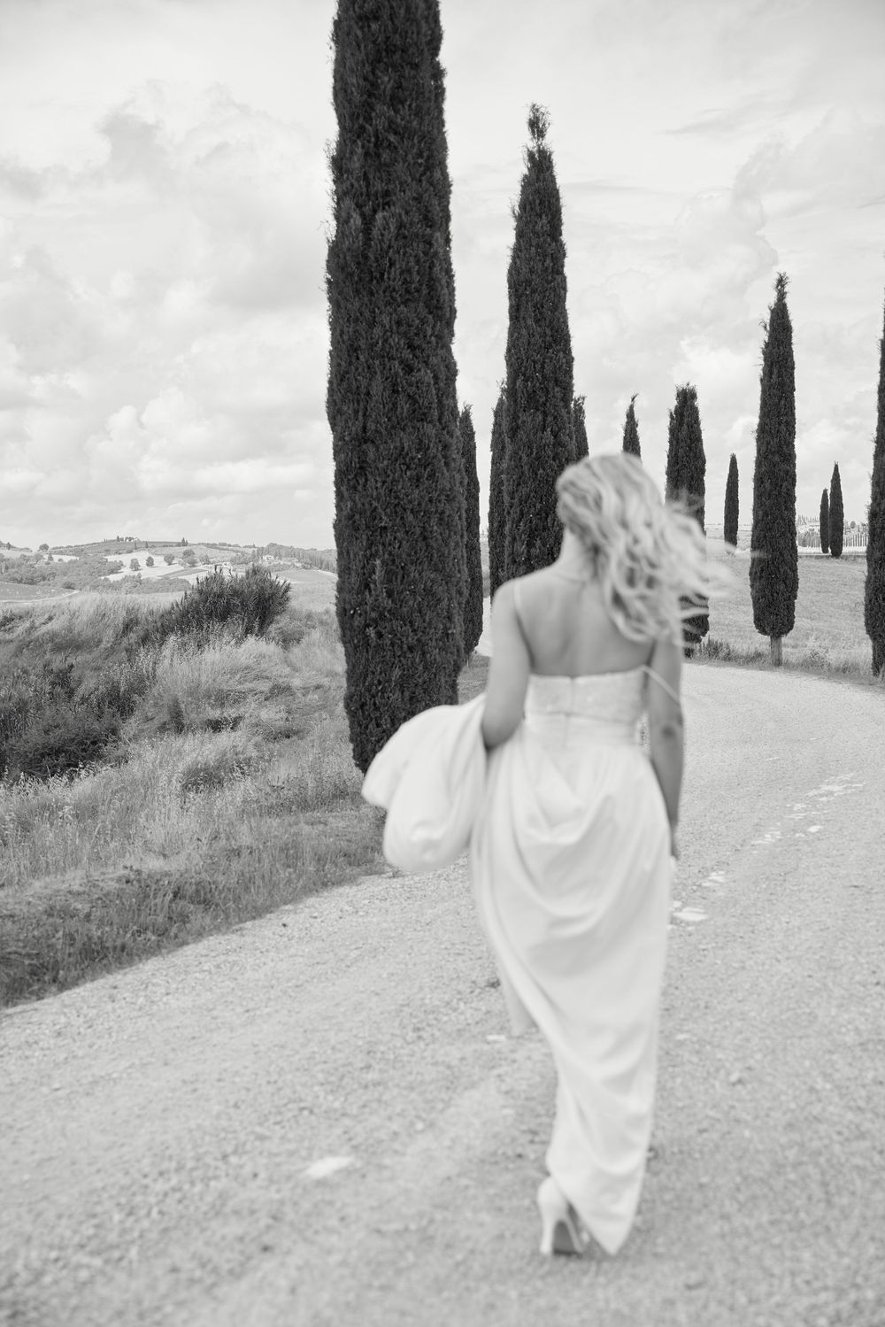 Black and white photo of a bride in a white gown walking down a cypress-lined road in Tuscany.