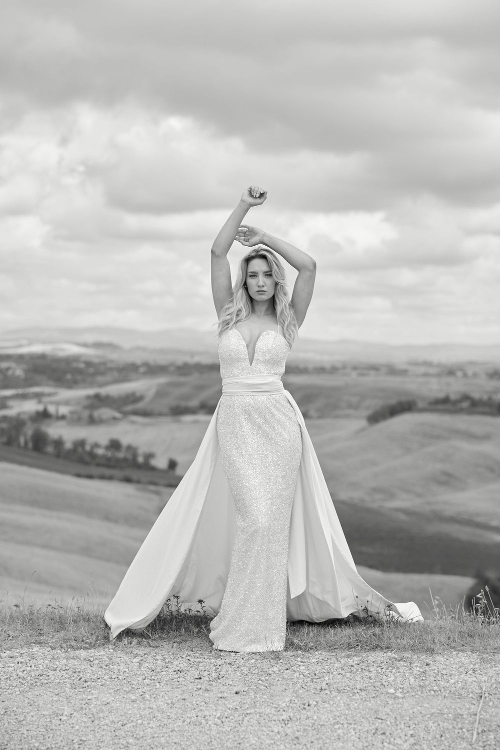 Elegant bride in flowing white gown poses with arms raised against scenic Tuscan countryside landscape.
