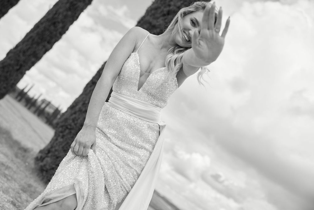 Smiling blonde bride in elegant V-neck wedding gown poses outdoors among trees in black and white photo.