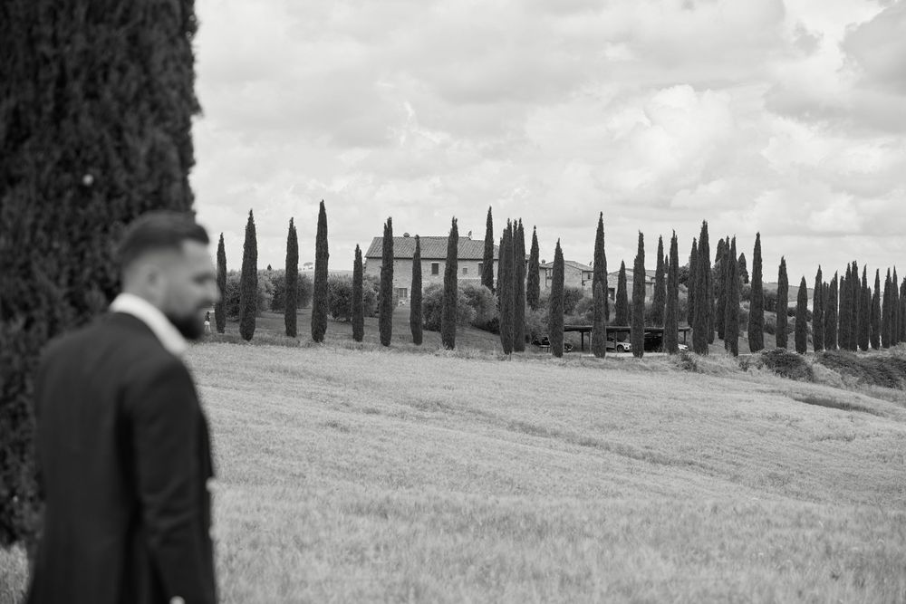 Black and white photo of a groom in a suit standing in a Tuscan field with cypress trees and villa backdrop.