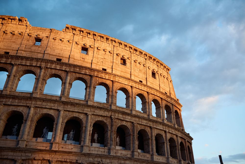 Golden sunset light illuminating ancient Roman Colosseum arches against dramatic sky.