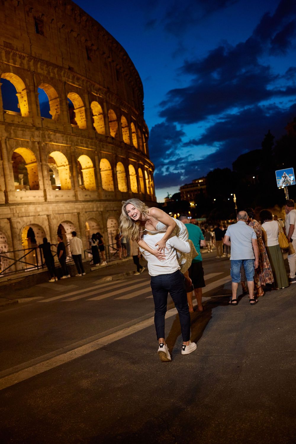 Joyful couple laughing as groom lifts bride near the beautifully lit Colosseum in Rome at dusk.