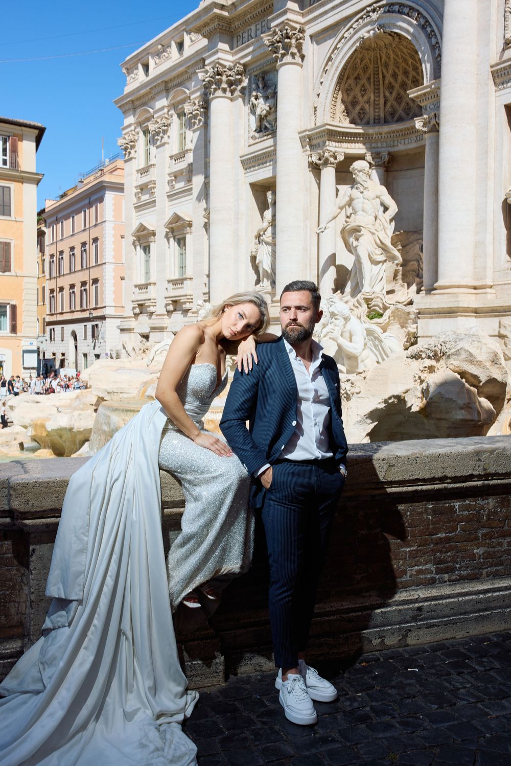 Elegant couple posing at Trevi Fountain in Rome, bride in silver gown and groom in navy suit.