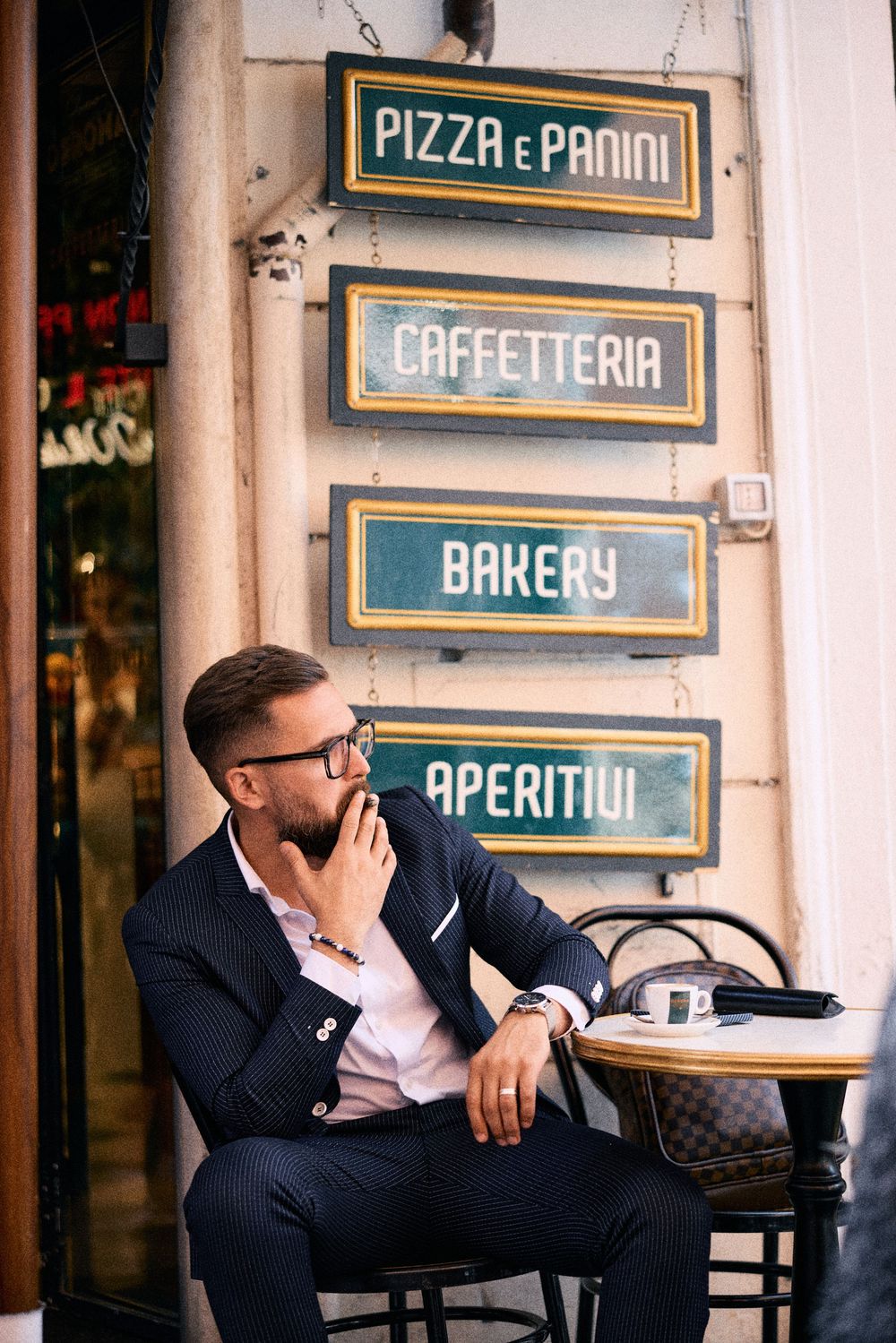 Stylish man in a navy suit sitting at an outdoor cafe table beneath vintage Italian restaurant signs.