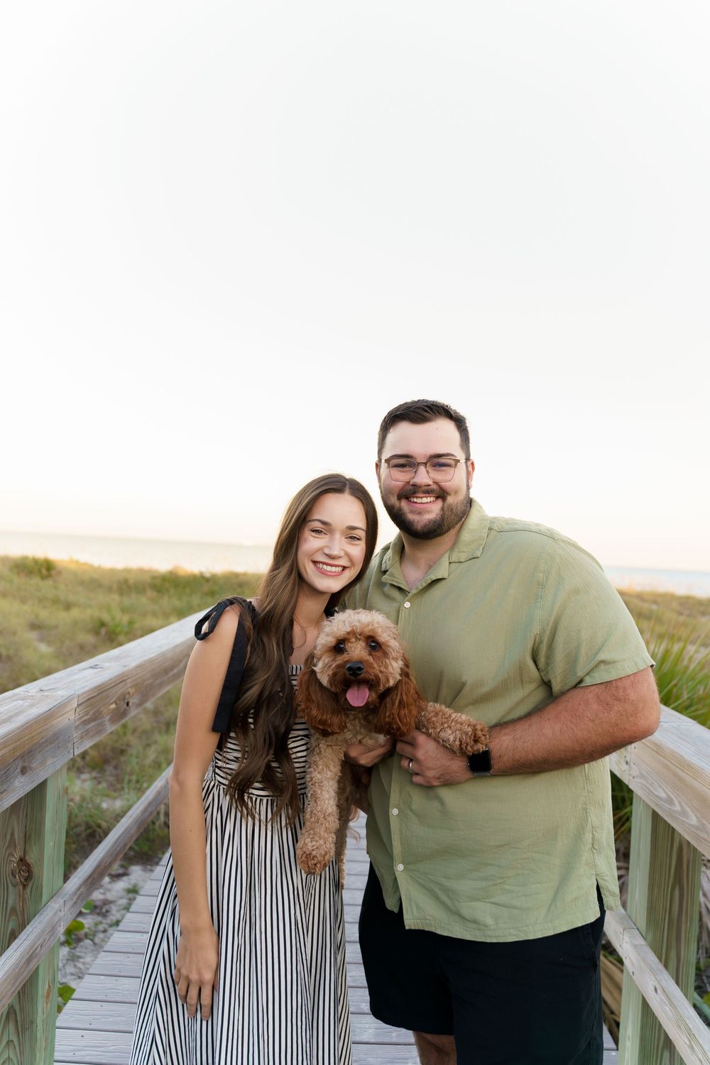A couple poses with their small brown dog on a wooden boardwalk at the beach during sunset.