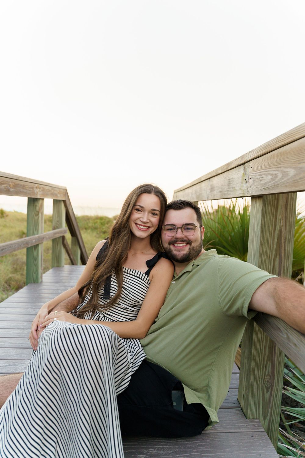 A couple sits together on a wooden boardwalk surrounded by coastal vegetation during sunset.