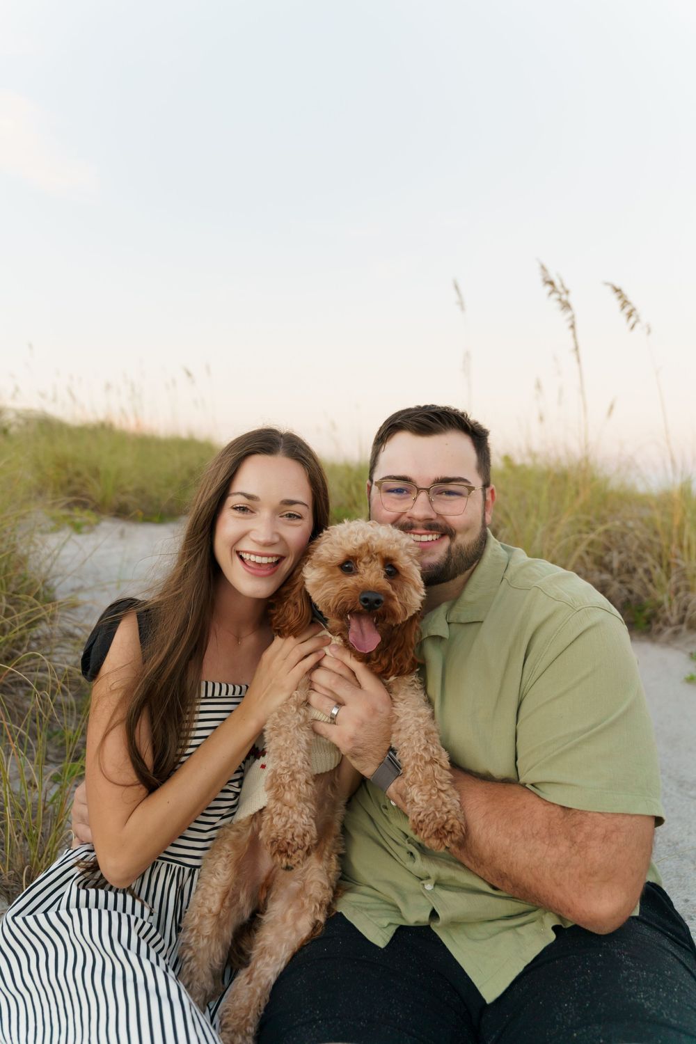 A couple and their goldendoodle dog share joyful moments together at the beach during sunset.