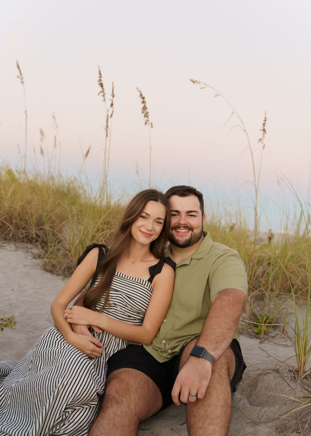 A couple sits together on a sandy beach with sea oats swaying in the foreground during golden hour.